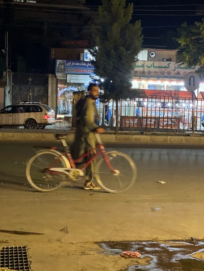 The image captures a nighttime scene on a street in Kabul, Afghanistan. A blurred figure on a red bicycle is the primary subject, depicted in motion. The cyclist wears a green garment and sandals. To the left, a white station wagon is parked. In the background, a building with signage in Arabic script, likely a shop or restaurant, is illuminated. The street appears wet, with puddles and debris visible on the asphalt. A metal grate is present in the foreground. The overall atmosphere is dimly lit, characteristic of an urban night environment.