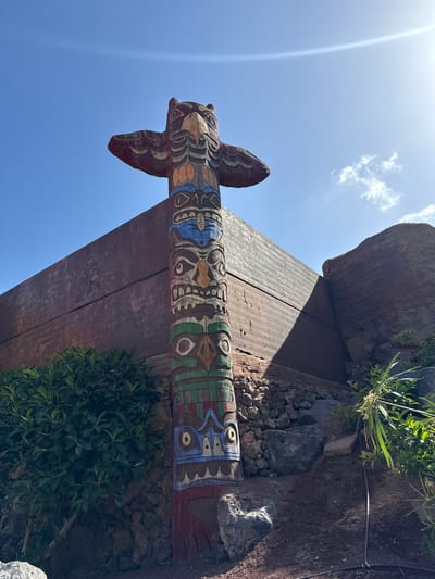 The image displays a brightly lit outdoor scene, captured under a clear blue sky with some wispy clouds and a prominent sun flare. The central subject is a tall, elaborately carved and vibrantly painted wooden totem pole, standing upright on the right side of the frame. The totem pole features stylized animal or anthropomorphic figures, with distinct eyes, mouths, and intricate geometric patterns in colors including blue, green, red, brown, orange, and white. Near its top, it has wing-like or arm-like protrusions.

The totem pole is positioned beneath a robust wooden archway or overhead structure, which extends horizontally across the middle of the image, casting a shadow in some areas. To the right of the totem pole, the ground is covered with dark, rough rocks and soil, interspersed with green leafy plants and bushes. Larger rocks and boulders are also visible, some appearing to be part of the natural landscape or decorative elements. The vegetation is varied, with some larger plants growing on top of the wooden structure and others near its base.

No people or activities are visible. The time of day is clearly bright daylight, likely midday or early afternoon, given the intensity of the sun and the shadows. The weather is sunny with a few scattered clouds. The rocky terrain and specific plant types hint at a dry, possibly volcanic environment, consistent with the location context of Tías, Spain. There is no visible text in the image.