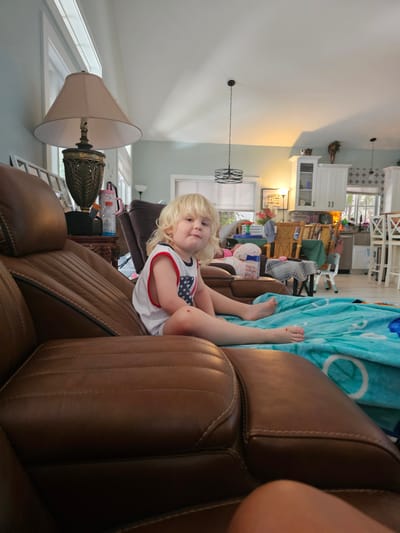 A blonde-haired toddler sits on a brown leather couch, facing the camera. The child wears a white tank top with red trim and a pattern of stars and stripes. The toddler's legs are propped up, and their left foot is visible, with a small red mark on the toe. Beside the toddler, a blue and white patterned blanket is draped over the couch.
The background reveals a living room and kitchen area. White cabinets and kitchen island are visible, along with a patterned backsplash. Furniture includes wooden chairs and tables, suggesting a family gathering or a well-lived-in home. A floor lamp with a tan shade stands to the left of the couch, casting a warm light. Another light fixture hangs from the ceiling. Various objects are scattered in the background, including bottles and what appears to be a child's toy. The overall impression is a casual, domestic setting. The lighting suggests it is daytime. The location is identified as Islamorada, United States of America.