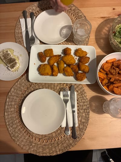 A table set for a meal features two place settings, each with a white plate on a woven placemat. One setting includes a small plate of seasoned feta cheese. A serving platter holds fried chicken bites. Bowls of salad and roasted sweet potatoes are shared. Glasses of water, one with ice, are also present. Silverware rests on white napkins. A hand is visible, possibly pouring a drink.