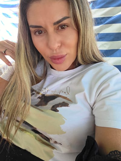The image is a close-up selfie of a woman with long, blonde hair and light brown eyes. She is wearing a white t-shirt with a graphic print. She has light makeup on, including lipstick. Her left hand is visible, resting on her hair, and she is wearing a ring on her left ring finger. The background consists of blue and white striped fabric, possibly a beach umbrella or chair. The lighting suggests it is daytime, likely outdoors. There is no information in the image to indicate a specific location in Ravnogor, Bulgaria.