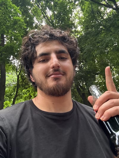 The media file is a selfie taken outdoors with a person holding a dark bottle and pointing at the camera. The person has dark, curly hair and a beard. They are wearing a black t-shirt. The background is filled with lush green trees, suggesting a natural setting. Sunlight filters through the leaves, indicating it is daytime. The person's facial expression is neutral, and their gesture with the bottle could imply a playful or casual interaction. The presence of trees suggests an outdoor location, possibly a park or a wooded area, consistent with the provided location context of Straszyn, Poland. There is no visible text in the image.