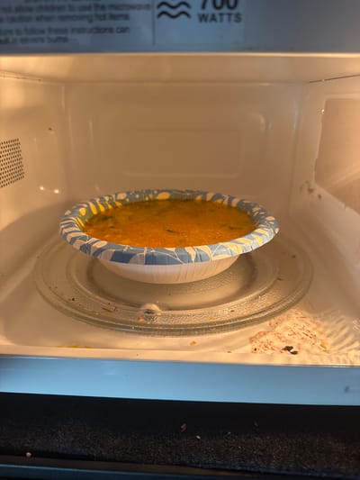 The image shows the inside of a microwave oven. A plate of food, likely a casserole or pasta dish, is centered on the glass turntable. The plate is white with a blue and yellow decorative rim. The turntable is clean except for some scattered crumbs and what appear to be small bits of food, possibly from previous use. The interior walls of the microwave are light-colored, and there are some dried splatters of food visible on the top and sides, indicating it needs cleaning. A label with text is visible on the left side of the microwave, but most of it is out of focus and cropped. The visible text includes "700 WATTS" and some instructions in smaller print. The lighting suggests the microwave is in use or has recently been used. The overall scene depicts a common household appliance with food inside, suggesting a meal is being prepared or reheated. There are no people or animals visible in the image. The setting is an interior domestic environment, likely a kitchen.
