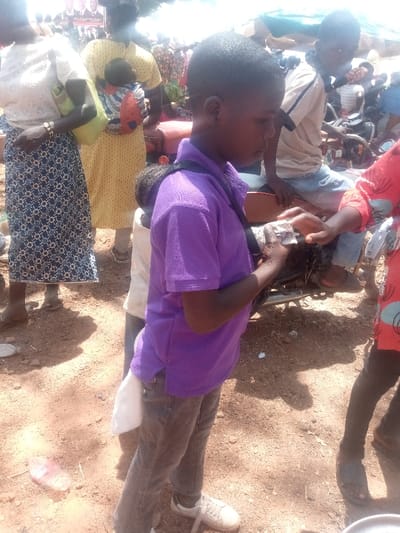 The image displays an outdoor, crowded scene in Kagoma, Nigeria, characterized by reddish-brown dirt ground with visible debris including a pink plastic bottle. The lighting suggests daytime.
In the foreground, a young male subject with short, dark hair, wearing a purple polo shirt, dark jeans, and white sneakers, stands facing left. A dark strap crosses his left shoulder. His right hand holds a small, light-colored, possibly paper-wrapped object. He is engaged in an exchange with another hand, partially visible on the far right, belonging to an individual wearing a red garment with light circular patterns. A smaller child in a white shirt is partially obscured behind the primary subject.
To the right of the central interaction, another male subject, possibly a youth, is seated on a dark-colored motorcycle, wearing a light-colored, striped short-sleeved shirt and blue jeans.
In the mid-ground on the left, an adult female stands, wearing a white, textured short-sleeved top and a long, dark blue skirt with a light geometric pattern. She carries a light green shoulder bag. Behind her, another adult female, facing away, wears a yellow polka-dot dress and carries an infant on her back in an orange-patterned baby carrier. Both women wear sandals.
The background is densely populated with numerous indistinct figures, suggesting a large gathering. Sections of light-colored tarpaulin or fabric awnings are visible overhead, along with a banner featuring multiple faces.