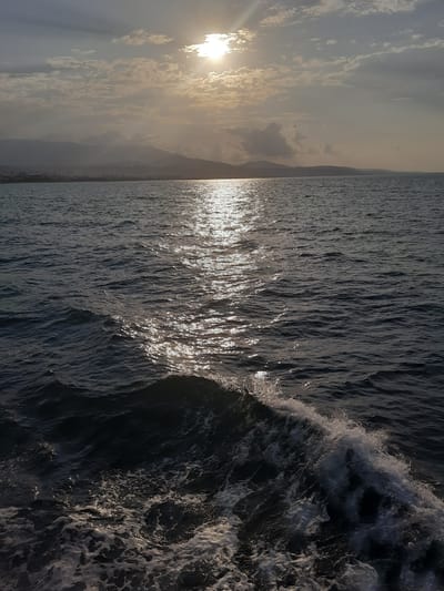The image shows a seascape with a shoreline and mountains in the distance. The sun is visible in the sky, partially obscured by clouds, casting a bright reflection on the water. The water is choppy, with waves and foam visible in the foreground. The shoreline appears to be a city with buildings and some vegetation. The overall impression is one of a natural landscape meeting an urban environment. The image is likely taken from a boat or ferry at sea. The specific location is not identifiable from the image alone, but the prompt indicates it is the city of Samsun, Turkey.