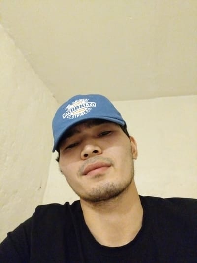A young man wearing a blue baseball cap with the text "THE ORIGINAL BROOKLYN CLOTHING CO." is pictured from the chest up. He has dark hair and is wearing a black t-shirt. The background is a plain, light-colored wall. The image appears to be taken indoors, possibly in a residential setting in the city of Tuzdybastau, Kazakhstan, as indicated by external information not present in the image itself.