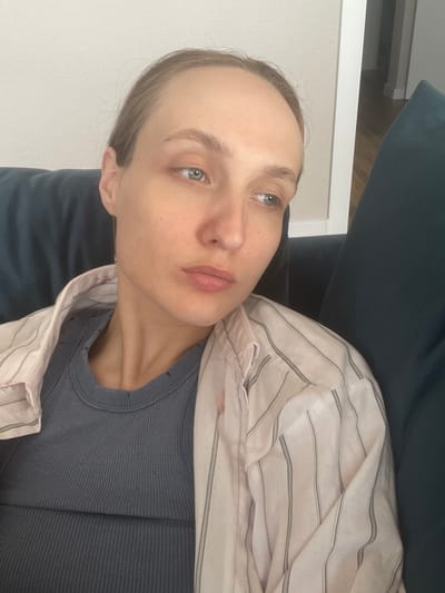 A pale-skinned individual with light brown hair and blue eyes lies in an indeterminate location, possibly indoors. The person wears a dark grey ribbed shirt and a light-colored, striped button-down shirt. The individual is partially obscured by what appears to be a dark blue sofa. The background consists of a white wall. The image is oriented sideways. There is no indication of the location being Sepych, Russia.