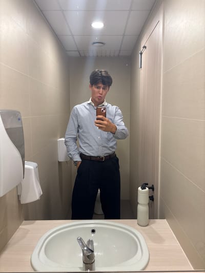 The media file shows a young man taking a selfie in a public restroom. The man is wearing a light blue and white striped long-sleeved shirt, black pants, and a brown belt. He is holding a smartphone in his right hand, angled to capture his reflection. His left hand is in his pocket. He has dark hair styled neatly. He appears to be looking at his phone with a neutral expression.

The restroom is tiled in light beige on the walls and floor. There is a white sink with a chrome faucet on the right side of the frame. Above the sink, a roll of toilet paper is mounted on a holder. In the background, there are white soap dispensers mounted on the wall. The lighting is bright and appears to be artificial, suggesting an indoor setting. The overall impression is that of a clean, modern public restroom. There are no visible windows or any indications of the time of day or weather outside. The location context provided is Basiglio, Italy.

The activity is a selfie taken by the subject in a restroom. No specific event is taking place, other than the act of taking a photograph. The notable details are the attire of the subject, the clean restroom environment, and the reflection of the subject in what appears to be a large mirror. There is no visible text in the image.