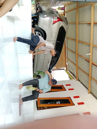 The image shows two barefoot men in what appears to be a driveway in Gampongjawa, Indonesia. One man, wearing dark pants and a light-colored shirt, has one hand on the side of a silver SUV parked in the driveway. The other man, wearing dark pants, a light blue shirt, and a baseball cap, is holding a plastic bag in one hand and what appears to be a pair of scissors in the other. The driveway is tiled, and the house has windows with wooden frames. The car is parked next to a structure with corrugated metal walls and wooden supports.