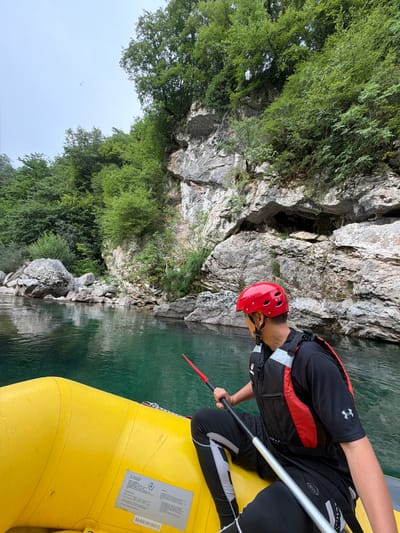 This is a medium shot, taken from a raft, looking up a river canyon. The main subject is a person in a raft, wearing a red helmet and a life vest over black athletic wear. The person is holding a paddle, with the red tip visible. The raft is bright yellow and has a sticker with text on its side. The river is a clear, green color, reflecting the rocky canyon walls. Lush green trees and vegetation cover the steep canyon sides. The time of day appears to be daytime, with overcast weather indicated by the bright, diffused light and the sky visible in the upper left. The scene evokes a sense of adventure and outdoor recreation, likely a whitewater rafting or kayaking trip. The location is Džajići, Bosnia and Herzegovina.

Visible text on the sticker on the raft includes: "PARAGON" and "BA-RIS19015H312". There are also smaller blocks of text that are difficult to read clearly in the image.
