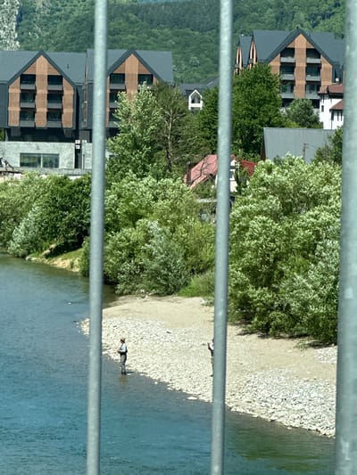 The image shows a wide river in the foreground, with a pebbly shore on the right side. Two individuals are fishing in the river. In the background, there are trees and modern buildings with dark roofs and wooden facades. The scene appears to be in Dulovine, Montenegro, characterized by its mountainous terrain and proximity to a river.