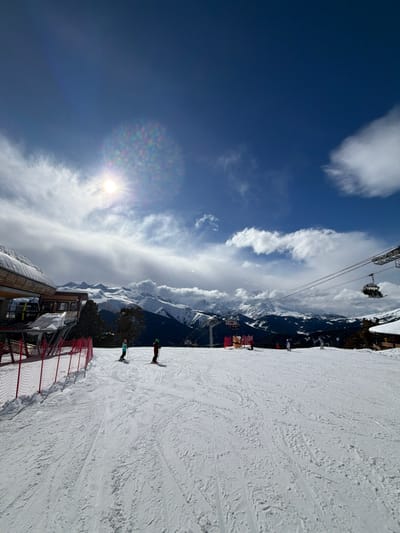 The image depicts a snow-covered ski resort identified as Lunnaya Polyana, Russia, under a clear blue sky with scattered white clouds. The sun is visible in the upper left quadrant, producing a lens flare. The foreground and midground consist of a wide ski slope marked with numerous ski tracks. On the left, a modern ski lift station with a multi-toned wooden roof and large windows is partially visible, flanked by a red mesh safety fence extending along the slope. Two individuals, equipped with skis and poles, are actively descending the slope in the midground left; one wears a light blue jacket, and the other a dark green jacket. To the right, a ski lift system spans the area, with multiple chairlifts, some occupied, visible on the cables. Further down the slope on the right, several other individuals are present, engaged in skiing or snowboarding near yellow and red padded obstacles. A small building with a wooden exterior is partially visible on the far right. The background features an extensive range of snow-capped mountains, partially covered by evergreen forests.