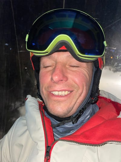 An adult male is depicted in a close-up, chest-up shot within an environment consistent with a ski resort in Jahorina, Bosnia and Herzegovina. The male, with fair skin, short stubble, and rosy cheeks, has his eyes closed and is smiling broadly, exposing his teeth. He is wearing winter sports attire, including a black ski helmet with red accents, a pair of lime-green framed ski goggles with blue reflective lenses pushed up onto the helmet, a patterned neck gaiter, and an off-white/light gray ski jacket featuring a red inner collar and a red main zipper. A black plastic buckle from the helmet strap is visible beneath his chin. The background is predominantly dark, with blurred yellow/orange lights and a textured, snow-like surface visible to the right, suggesting a nighttime or low-light setting on a ski slope or within a ski lift.