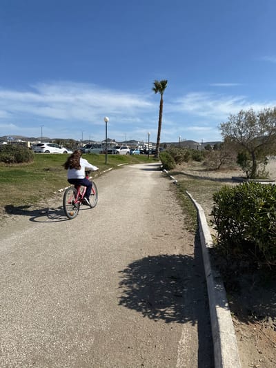 The image depicts an outdoor scene on a bright, sunny day in Anatoliki Attiki, Greece. The main subject is a person with long, dark hair, wearing a white long-sleeved top and dark trousers, riding or paused on a red bicycle. The person and bike are on a light-colored paved path that curves slightly. To the right of the path (when viewed upright), there is a grassy verge bordered by a concrete curb, and a large, dense shadow from a nearby tree falls across the path.

In the midground, to the left of the path, a grassy area separates it from a parking lot where several white and light-colored cars are visible. Multiple lampposts with spherical tops are spaced along this area. A tall, slender palm tree stands prominently above the parking lot, reaching towards the clear blue sky, which has a few wispy clouds. In the far background, distant hills or mountains with sparse vegetation and some scattered buildings are visible. The strong shadows cast by the tree and bicycle indicate bright sunlight, suggesting a time between mid-morning and late afternoon. There is no visible text in the image.