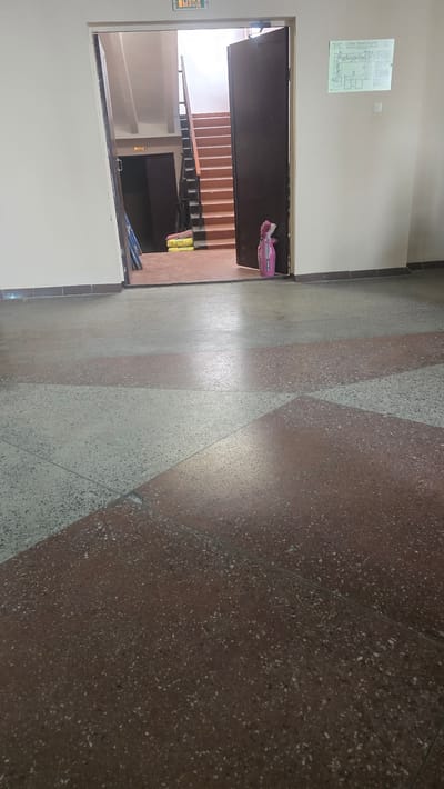 The image depicts a hallway with a staircase visible through an open door. The hallway floor is a terrazzo with a pattern of red and light gray diamond shapes. To the left, a door is open, revealing a staircase with light brown steps and dark railings. A pink duffel bag rests on the floor near the open door, and two yellow bags of what appear to be building materials are placed near the bottom of the stairs.
On the wall to the left of the doorway, a white sign with a diagram is mounted. There is also a green sign with text that appears to say "ВЫХОД" (EXIT) in Russian, mounted on the wall to the left of the doorway. Another similar green sign is visible on the wall inside the hallway leading to another door.
The lighting suggests it is daytime, possibly indoors with artificial lighting. The scene appears to be a public building, such as an apartment complex or an office building, given the presence of a staircase and exit signs. There are no people visible in the image. The overall impression is a utilitarian and somewhat dated interior space.