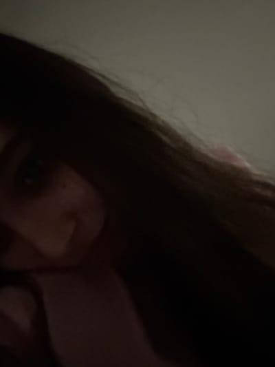 A dimly lit, close-up shot focuses on a person's face, partially obscured by dark hair. One eye is visible, and a hint of nose and mouth are suggested. The background consists of a plain, lighter-colored surface. The overall image has a slightly grainy texture, possibly due to low-light conditions.