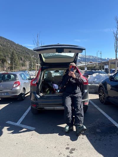 The image depicts an outdoor parking lot in La Salle-les-Alpes, France, under a clear blue sky. A person, wearing a black ski jacket, black ski pants, and greenish-black ski boots, is seated on the open rear bumper of a dark-colored SUV, likely a Volvo. The individual's face is largely obscured by reddish-orange ski goggles and a beanie, while they hold a smartphone in both hands at chest level, seemingly operating its camera. The SUV's tailgate is fully open, revealing ski equipment, including what appears to be ski poles and two helmets (one purple, one black), within the cargo area.
To the left of the SUV, a silver Renault Clio with French license plate "600-TA" is parked in an adjacent space. A portion of another dark vehicle is visible to the right. The ground is asphalt with white parking lines, and shadows are cast by the person and vehicles, indicating direct sunlight. In the background, a steep mountain slope covered with evergreen trees rises, interspersed with bare deciduous trees. Patches of snow are visible on the ground beyond the paved area. Distant snow-capped mountains and buildings are visible further in the background.