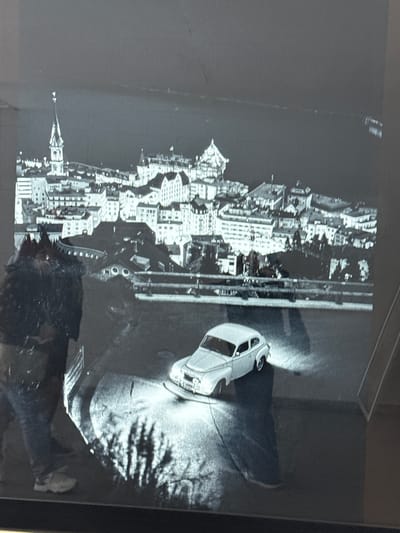 A black and white photograph depicts a cityscape at night with a lake or body of water in the background. In the foreground, a vintage white car is parked on a paved surface, illuminated by its headlights which cast bright beams onto the ground and a shadow of foliage. To the left of the car, a person wearing jeans and sneakers stands, their back partially to the camera, holding what appears to be a bag. The city skyline features numerous buildings with pitched roofs, and a prominent tower with a spire rises above them. The overall scene suggests a quiet, possibly historic, urban environment at night. The specific location within Sesto San Giovanni, Italy, cannot be determined from the visual information provided.