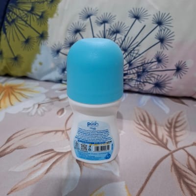 A close-up, slightly angled shot shows a Posh Hijab anti-perspirant deodorant roll-on bottle with a bright blue cap. The bottle is white and has text printed on it in multiple languages. The product claims to keep users cool and refreshed with a fragrance that lasts all day. It also states that it contains 99% anti-bacteria and 0% alcohol. The bottle is placed on a fabric surface with a floral pattern. The background consists of a textured fabric with large, stylized dandelion-like motifs in navy blue and yellow on the left, and similar motifs in navy blue and pink on the right. The lighting is bright and appears to be natural. The image does not provide any information about the location in Reudeuep, Indonesia.