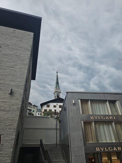 The image shows a low-angle view of buildings in St. Moritz, Switzerland, under a cloudy sky. In the foreground on the left, a modern building with a grey stone facade and a dark roof is visible. To the right, another modern building with a grey facade features large windows with patterned curtains and two prominent "BVLGARI" signs in gold lettering. Behind these buildings, a historic church tower with a green spire and a clock face rises into the sky. Other buildings, including residential structures, are visible on a slope in the background. There are no people or animals visible in the image. The scene depicts an urban environment with a mix of contemporary and historical architecture.