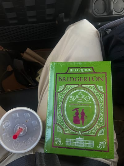 The image shows a book titled "Bridgerton: Daphne & Anthony" by Julia Quinn resting on a person's lap, likely inside a vehicle. A cup with a red straw is beside the book. The specific location in Comfort, United States of America is not evident in the image.