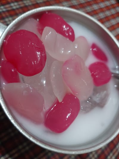 A close-up, overhead view shows a metallic bowl filled with a white liquid and several gelatinous, oval-shaped pieces. Some of the pieces are bright pink, while others are translucent white or pale pink. Ice cubes are visible beneath the pieces and the liquid. The bowl is positioned on a surface covered with a red and white checkered cloth. The scene is well-lit, with reflections visible on the surface of the gelatinous pieces and the liquid. The image does not provide specific details about the location being in Jelutung, Indonesia.