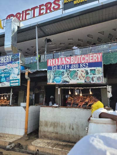 This image captures an active street-side commercial area in Kimorori, Kenya, centered around a butchery and another establishment. The most prominent business is "ROJAS BUTCHERY," identified by a large vertical sign displaying its name, phone number (Tel: 0719 480 852), and images of cooked dishes. Below this, another sign indicates available payment methods: M-PESA, KCB BANK, and EQUITY.
To the right of the butchery sign, an outdoor grilling station is in full operation, where several pieces of skewered meat, typical of Kenyan nyama choma, are being roasted over open flames. Bright lights illuminate the grilling area. A person in a yellow shirt is actively tending to the grill. On the far right, another individual, partially obscured by a large white bucket they are carrying, is present. Inside the butchery, a person wearing a black cap or hoodie is visible.
To the left, a large horizontal sign, partially visible, reads "DRIFTERS" (with "TFITTERS" and "S" being clear) and also mentions "ACCU MUSIC," suggesting an entertainment or music-related venue. The buildings feature a metal roof overhang and visible upper-story architecture with glass railings. A silver ventilation duct extends from one of the structures. The ground level consists of concrete and tiled surfaces. The overall scene suggests a busy daytime or early evening setting with local commerce.