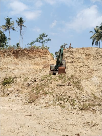 A wide shot shows an excavator situated on a large, excavated dirt embankment under a blue sky with scattered clouds. The embankment is composed of layered soil and rock, with some sparse vegetation growing on its surface and at its base. Several palm trees are visible on the ridge of the embankment, with one on the far left, two in the center left, and another on the far right. The excavator, a green machine with a large, rusty bucket, is positioned in the middle of the embankment, appearing to be stationary. The ground in the foreground is rough and sandy with scattered rocks and small plants. The scene appears to be a construction or excavation site.
