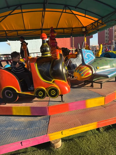 This outdoor, brightly lit image captures a segment of a children's amusement park ride, likely located in Ait Ouallal, Morocco. The scene features a colorful track with several ride vehicles and a young child as the primary subject.
In the foreground, a young child with dark, curly hair, wearing a striped long-sleeved shirt, sits in an orange and black ride vehicle with yellow accents. This vehicle has a futuristic or robotic design, featuring prominent circular light-like elements at the front. The child is looking to the left, with one hand near their mouth, suggesting a moment of observation or anticipation. Further along the track, other ride vehicles are visible, including a green and white one with a striped snout, possibly resembling a fish or alien creature, and additional animal-shaped vehicles (like a giraffe and a horse) under a canopy.
The ride track consists of textured metal sections, interspersed with vibrant, painted barriers in shades of yellow, pink, and orange. To the left, a large, arched canopy or awning, with a bright yellow fabric top and a green metal frame, provides shade over part of the ride. On the right side of the track, a patch of artificial green grass is visible. In the background, under a clear blue sky, brick-colored buildings can be seen.
The activity depicted is likely children enjoying or waiting for a ride at an amusement park or fair. The bright sunlight and distinct shadows indicate it is daytime. No discernible text is visible within the image.