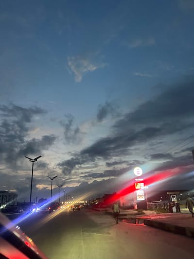 The image captures a street scene in Lagos, Nigeria, at dusk. The sky is filled with dramatic, cloudy formations, painted with the subdued colors of twilight. A row of tall streetlights lines the road, casting pools of light as the day ends. On the right side of the street, a Quickmart gas station is visible, its signage illuminated by bright red and white lights, displaying prices that are blurred by the light's intensity. A few cars are present on the road, their headlights and taillights creating streaks of color due to the camera's exposure. Two figures are walking on the side of the road, one closer to the foreground and another further down the street. The overall atmosphere is one of a busy urban environment transitioning into evening.