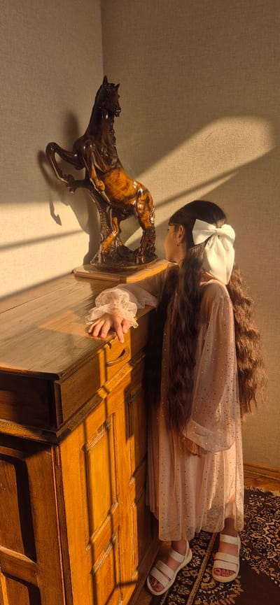 A young girl with very long, dark hair tied with a large white bow leans over a wooden dresser. She wears a light pink, sparkly dress and white sandals. Her hands are on the dresser, and she appears to be looking at a wooden sculpture of a horse. The sculpture is dark brown and appears to be in motion. The scene is lit by sunlight streaming in from the left, casting shadows across the dresser and the wall. The wall appears to be textured, possibly wallpaper or a painted finish. A patterned rug is visible on the right side of the image. The time of day is likely afternoon due to the angle of the sun and the long shadows. The setting appears to be indoors, in a room with wooden furniture and a textured wall. The girl's posture suggests curiosity or contemplation. There is no visible text in the image.