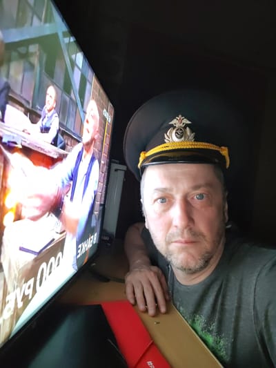A man is shown in a selfie indoors, next to a television screen. The man is wearing a dark peaked cap with a metallic insignia and gold rope trim. He has a beard and light-colored eyes. The television screen shows a scene with two people. One person is looking up with an expression of surprise, while the other person is partially visible on the left. Text is visible on the television screen, written in a script that appears to be Cyrillic. The man is leaning on a wooden surface next to the television, with a red object visible below. The environment is dimly lit. Although the city of Ermolino, Russia is mentioned, there is no visual evidence in the image to confirm the location.