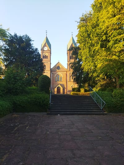 The image shows a brick church with two towers and a green roof, located in Leverkusen, Germany. The church is surrounded by trees and lush greenery. In front of the church, there is a set of dark stone stairs leading up to the entrance. A stone pathway is visible in the foreground. The sky is clear and blue, and the sun is casting a warm glow on the scene, suggesting it is either early morning or late afternoon. There are no people visible in the image. The overall impression is one of peace and tranquility.