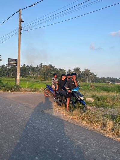 Three young men are posing for a photo on a blue motorcycle parked on the side of a paved road. The man in the front is wearing a black t-shirt and black shorts, and he is giving a peace sign with his fingers. The man behind him is wearing a black t-shirt and appears to be looking at the camera. The third man is wearing a black and white patterned shirt and is also looking towards the camera. The motorcycle is parked on a grassy embankment next to the road, with dry, tall grass and some green vegetation.

In the background, there is a lush green rice paddy, with several palm trees and other tropical foliage. In the distance, a few houses are visible. The sky is clear and blue with a few wispy clouds. The lighting suggests it is late afternoon or early evening, as the sun is casting long shadows. There is a power pole with wires running across the left side of the frame. A sign on a post near the road reads "Lombok Hidden Villa". The overall scene suggests a casual gathering of friends in a rural, tropical setting in Indonesia. The men appear relaxed and happy.