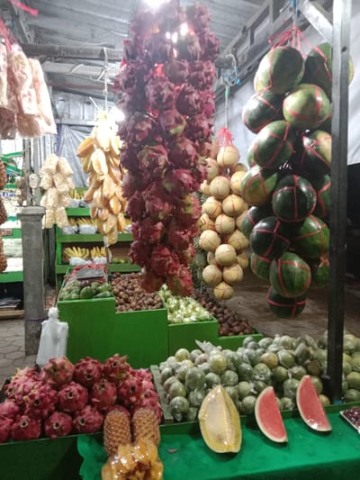 The image depicts a fruit and snack stall within a market in Sekopek Wetan, Indonesia. The scene is illuminated by overhead artificial lighting, suggesting a covered or indoor setting with a corrugated metal ceiling and visible structural beams.
Various produce items are suspended from the ceiling and overhead structures:
* On the far left, multiple bundles of light-colored, crunchy snacks are hanging.
* Next to these, three elongated clusters of yellow star fruit (carambola) are displayed.
* Centrally, a dense, vertical column of red dragon fruit (pitaya) is suspended.
* To its right, several bundles of light-toned, spherical fruits, possibly small melons, are hanging, each secured with string.
* On the far right, a vertical arrangement of green watermelons, each individually secured with red strapping, hangs from a dark metal pole.
Below the hanging produce, a multi-tiered green display unit occupies the foreground and mid-ground. On its lower shelves, a large mound of fresh red dragon fruit is visible, alongside two whole pineapples and several slices of cut fruit: one half of a yellow melon and two slices of red watermelon. Further back on the green shelving, distinct sections display various fruits: green avocados, dark red mangosteen, light green guavas (some individually wrapped in plastic), and brown salak (snake fruit). A weighing scale is positioned on one of the green display sections containing avocados. In the background, additional market shelving holds bananas and other obscured produce. The ground appears to be a paved surface. No individuals are present within the frame.
