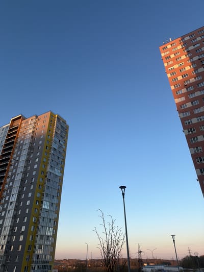 The image shows a view looking upward in Анкудиновка, Russia. At the top is a tall, salmon-colored building with a grid of windows. At the bottom is another tall building, painted with yellow and gray and also featuring a grid of windows. In the background, the sky is blue near the buildings and fades into a muted orange-beige near the horizon. Street lamps and bare tree branches are visible in the mid-ground, against the sky. Power lines and what appear to be buildings further in the distance can be seen.