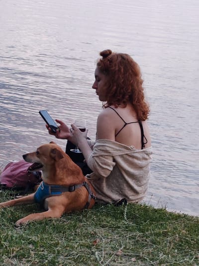 A young woman with curly red hair, styled in a bun, sits on the grassy shore of a body of water. She is wearing a light-colored off-the-shoulder top over a black spaghetti strap top. In her right hand, she holds a stemmed glass containing a dark liquid, likely wine. Her left hand holds a dark-colored smartphone. Beside her, a brown dog wearing a blue harness lies on the grass, looking to the left. A pink bag is visible behind the dog. The water is calm with ripples reflecting the sky. The scene appears to be in a natural setting, possibly a park or lakefront. The location is identified as Нарацкі сельскі Савет, Belarus.