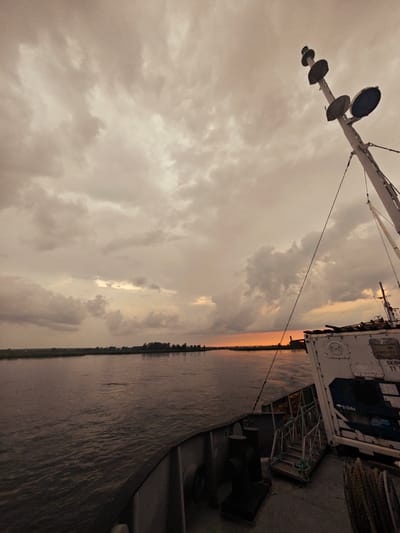 The image depicts a view from the deck of a vessel sailing on a wide river, likely the Yenisei River, near Kazachinskoye, Russia, during sunset. The sky is filled with overcast, grey clouds, with a hint of orange light peeking through on the horizon, suggesting the sun is setting. The water of the river is dark and calm, reflecting the cloudy sky. On the right side of the image, the deck of the ship is visible, showing railings, ropes, and a white container with markings. A tall mast with lights and antennas extends upwards on the right. The distant shore is lined with trees and appears flat. The overall atmosphere is somber and serene, characteristic of a cloudy sunset over a large body of water. There are no people visible in the image.