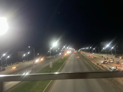 The image shows an aerial view of a highway at night in Sauka, Nigeria. The highway is illuminated by streetlights, and there are several cars on the road, some of which appear to be in a state of disrepair or have been involved in an incident. There are also people standing around some of the vehicles. The overall scene suggests a potentially chaotic or unusual situation on the highway.