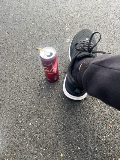 The image displays a close-up, top-down view of an outdoor paved surface. On the right side, a human left foot is visible, clad in a black athletic shoe with a white sole and black laces, worn over a black sock. The lower portion of a dark-colored garment, possibly sweatpants, is visible around the ankle. To the left of the foot, a red aluminum can of "COMET Cola cherry" is positioned upright on the pavement. The can features a "VEGAN" logo and the text "PENNY," indicating its brand. A partially smoked hand-rolled cigarette or joint is inserted into the can's opened top. The ground is dark gray asphalt, containing small light-colored aggregate and sparse debris, such as tiny leaves and pebbles. The specific location in Herne, Germany, cannot be confirmed from the visual information provided.
