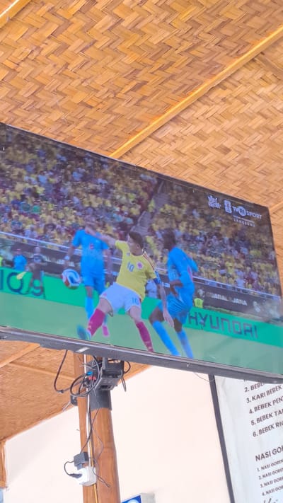 An outdoor television displays a soccer match. The television is mounted on a wooden pole and is viewed from below, looking upwards at an angle. The screen shows a close-up of a soccer game in progress, with players in yellow and blue uniforms. The background is a stadium filled with spectators. The television screen itself has logos and text displayed, including "TVRI SPORT" and "HYUNDAI". Above the television, a woven bamboo ceiling is visible. To the right of the television, a white sign with a menu is partially visible, listing food items such as "BEBEK" and "NASI GORENG." The scene appears to be an outdoor seating area, possibly a restaurant or cafe. The overall environment suggests a warm climate.