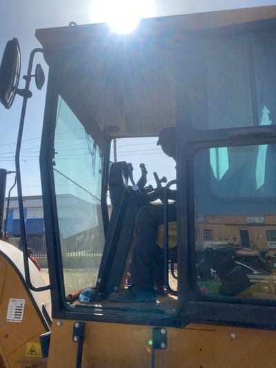 The image captures the interior of a heavy construction machinery cab in Rijiyar Lemo, Nigeria, under bright daylight conditions. The view is from outside the machine, looking into its left side. A male operator is seated inside, largely silhouetted against intense sun glare originating from the upper-center of the frame, which causes significant lens flare. He wears a cap and dark clothing, with his hands positioned near multiple control levers and a steering mechanism. A clear plastic water bottle lies on the cab floor. The machine's exterior is yellow, featuring a "SAVVY" decal and hazard symbols. Through the clear glass of the left window, a bright blue sky and a building with a blue facade are visible. A small, partially obscured figure (possibly a child in red clothing) is visible at ground level outside the machine on the left. The right cab window reflects an urban street scene, including a light-colored building with inverted text (e.g., "BOSSAS") indicative of commercial establishments, and indistinct figures of people, suggesting pedestrians and potential vehicles on a street. Side mirrors are attached to the exterior of the cab frame.