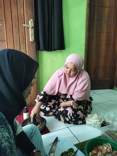Two women are captured inside a home, likely in Leuwiliang, Indonesia, engaged in food preparation. Both are seated on a white tiled floor, wearing hijabs.
On the left, partially visible from the back, a woman in a black hijab, glasses, and blue denim pants is grinding ingredients in a dark gray mortar with a pestle. Her watch is visible on her left wrist. To her right, a second woman, dressed in a light pink hijab and a black floral-patterned long-sleeved garment, sits facing forward but looking down, observing or handling food items.
Various cooking tools and ingredients surround them. A light blue chopping board with finely chopped green chilies or other vegetables and a knife rests on the floor. Adjacent to this, a green bowl holds what appears to be a salad or mixed chopped food. Several plastic containers are also present, one holding white crisps or crackers and another containing darker food. A white bottle or dispenser sits near the woman on the left.
The background features a bright green painted wall on the right and a wooden door with a metallic handle on the left. A piece of black fabric, possibly a curtain or hanging garment, covers part of the wall near the door. The scene depicts a focused and communal atmosphere of culinary activity. No visible text is present.
