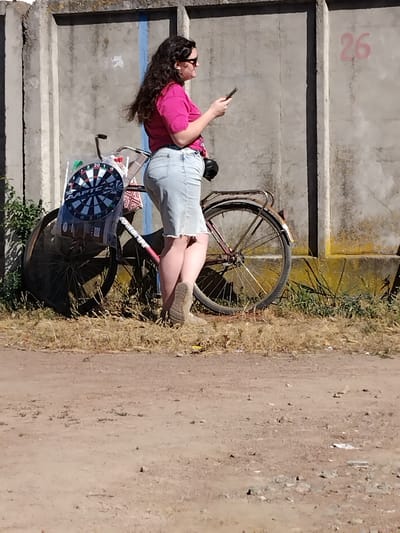 In Irshansk, Ukraine, a woman with long, dark, wavy hair and sunglasses stands beside a bicycle. She is wearing a pink short-sleeved shirt and a light blue denim skirt. The bicycle has a dartboard attached to its frame and a small red taillight. The woman is holding a mobile phone in her right hand, and her left hand is resting on the bicycle's frame. The scene takes place outdoors, with a concrete wall behind the woman and the bicycle, and dry grass and dirt in the foreground. The wall has the number "26" spray-painted in pink on its surface.