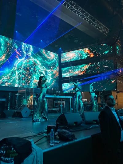 The image depicts a nighttime outdoor stage performance, possibly in Misaktsieli, Georgia. The stage is illuminated by bright blue laser lights and large LED screens displaying abstract, cosmic imagery of a human form and swirling galaxies. On the stage, four women in shimmering, sequined costumes are performing a dance. The woman in the foreground on the left is in a pose with her arms raised, while the others are positioned behind her, also dancing. In the background, musical equipment and speakers are visible, along with a man in a dark suit and white shirt standing to the right of the frame, seemingly observing the performance. The overall atmosphere is vibrant and energetic, characteristic of a live entertainment event.