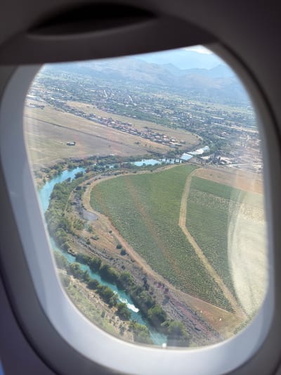 The image is a high-angle aerial view taken through an airplane window, overlooking the city of Donja Cijevna, Montenegro. The landscape features a winding, turquoise river with lush green vegetation along its banks. Adjacent to the river are fields of crops, appearing to be vineyards, with distinct rows of plants. Further inland, a settlement with red-tiled roofs is visible, interspersed with trees. In the background, rolling hills and mountains stretch towards the horizon under a hazy, light-blue sky. The scene is dominated by natural elements such as the river, vegetation, and mountains, alongside man-made structures including the village and agricultural fields. There are no visible people or specific actions depicted.