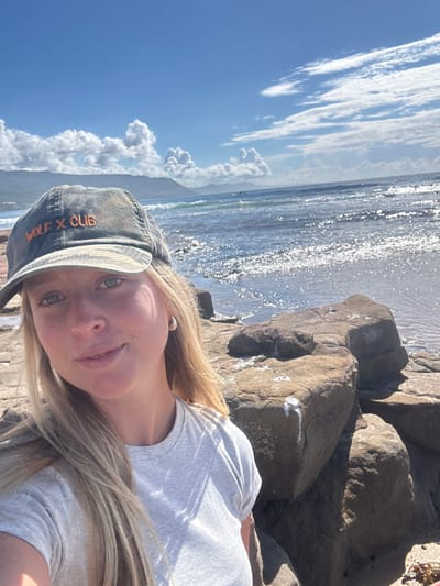 A female subject is captured in a selfie-style photograph on a rocky beach. She has fair skin, blonde hair, and blue eyes, and is wearing a light grey t-shirt and a distressed dark grey or green baseball cap with "WOLF X CLUB" embroidered in orange text. A small, light-colored hoop earring is visible in her left ear. She is looking directly at the camera with a slight smile.
The immediate foreground and midground feature prominent, irregular brown and grey rock formations, some exhibiting white residues consistent with dried sea spray or bird guano. Beyond the rocks, the Pacific Ocean extends with moderate waves, whitecaps, and significant glinting reflections from direct sunlight on the water surface. In the distant background, a green, mountainous headland or escarpment (consistent with the Illawarra Escarpment) runs parallel to the coastline. Several small, dark figures, likely surfers or swimmers, are visible in the distant ocean. The sky above is clear blue with scattered white cumulus clouds.
The location is identified as a rocky section of a beach, consistent with **Coledale Beach**, situated within the Northern Illawarra region of the **Wollongong City Council, Australia**.