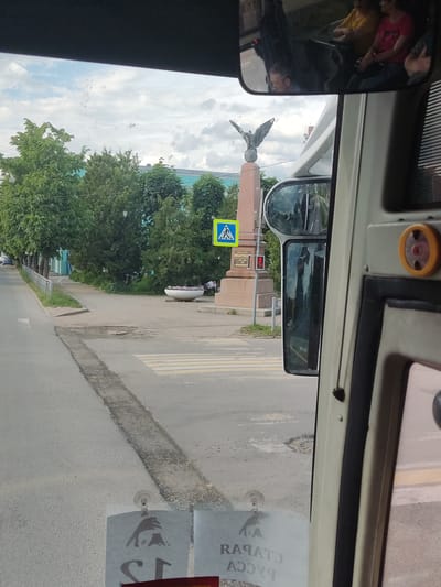The image provides a frontal view from inside a vehicle onto an urban street scene in Staraya Russa, Russia.

The scene depicts a street with an asphalt surface, marked by a pedestrian crossing with white zebra stripes in the mid-ground. A distinct rough linear crack or demarcation runs down the center of the visible road segment. To the left, a paved sidewalk is bordered by a white metal fence and a line of mature green trees. A light blue building is partially visible behind these trees in the distance.

On the right side of the frame, another paved area leads to a prominent monument. This monument consists of a tall, reddish-brown stone obelisk topped by a bronze eagle figure with outstretched wings, perched on a sphere. A yellow square pedestrian crossing sign, featuring a black walking person symbol, is visible just to the left of the monument's base. Further right, a red pedestrian traffic light is illuminated. Adjacent to the monument, a large, white, circular planter filled with colorful flowers is situated on the sidewalk, with several people visible sitting in the background nearby. The sky above is cloudy.

Inside the vehicle, the top portion of the dashboard or headliner is dark. The rearview mirror reflects at least three occupants: a man in the foreground (likely the driver or front passenger) and two women seated further back. A portion of the vehicle's right-side frame and a side mirror are visible. On the windshield, two transparent hangers with suction cups are present. One hanger displays a silhouette of an animal (resembling a wolf or dog) jumping, with mirrored text below it that reads "СТАРАЯ РУССА" (Staraya Russa).

The monument is identifiable as the Monument to the Defenders of the Fatherland (also known as the Eagle Monument), located in Staraya Russa, likely at or near the intersection of Ulitsa Volodarskogo and Ulitsa Karla Marksa.