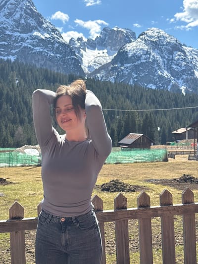 A medium shot presents a young woman with her hands behind her head, positioned in front of a weathered wooden fence. She is wearing a gray long-sleeved shirt and dark jeans. Beyond the fence stretches a landscape of grassy terrain dotted with what appears to be animal burrows. In the background, dense coniferous trees cover the hillside. In the distance, towering snow-capped mountains punctuate the scene, set against a blue sky with scattered clouds. The photograph was taken near Sappada / Plodn / Sapade, Italy.