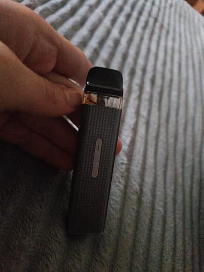 A close-up shot shows a hand holding a black vape pen. The vape pen has a textured surface and a metallic strip with a small button. The background consists of a gray, striped fabric. The image does not contain any information about the location in the city of Horlivka, Ukraine.
